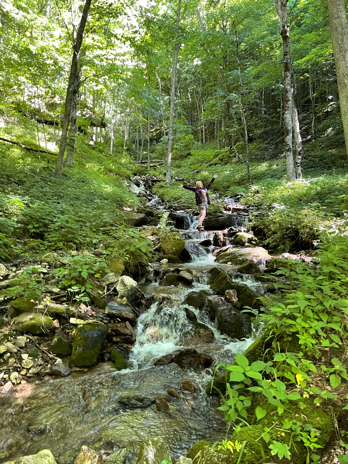 Karen standing arms wide beside a mountain creek in the forest